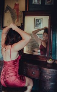A woman in a red dress looks in a vintage mirror while adjusting her hair, in a classic room.